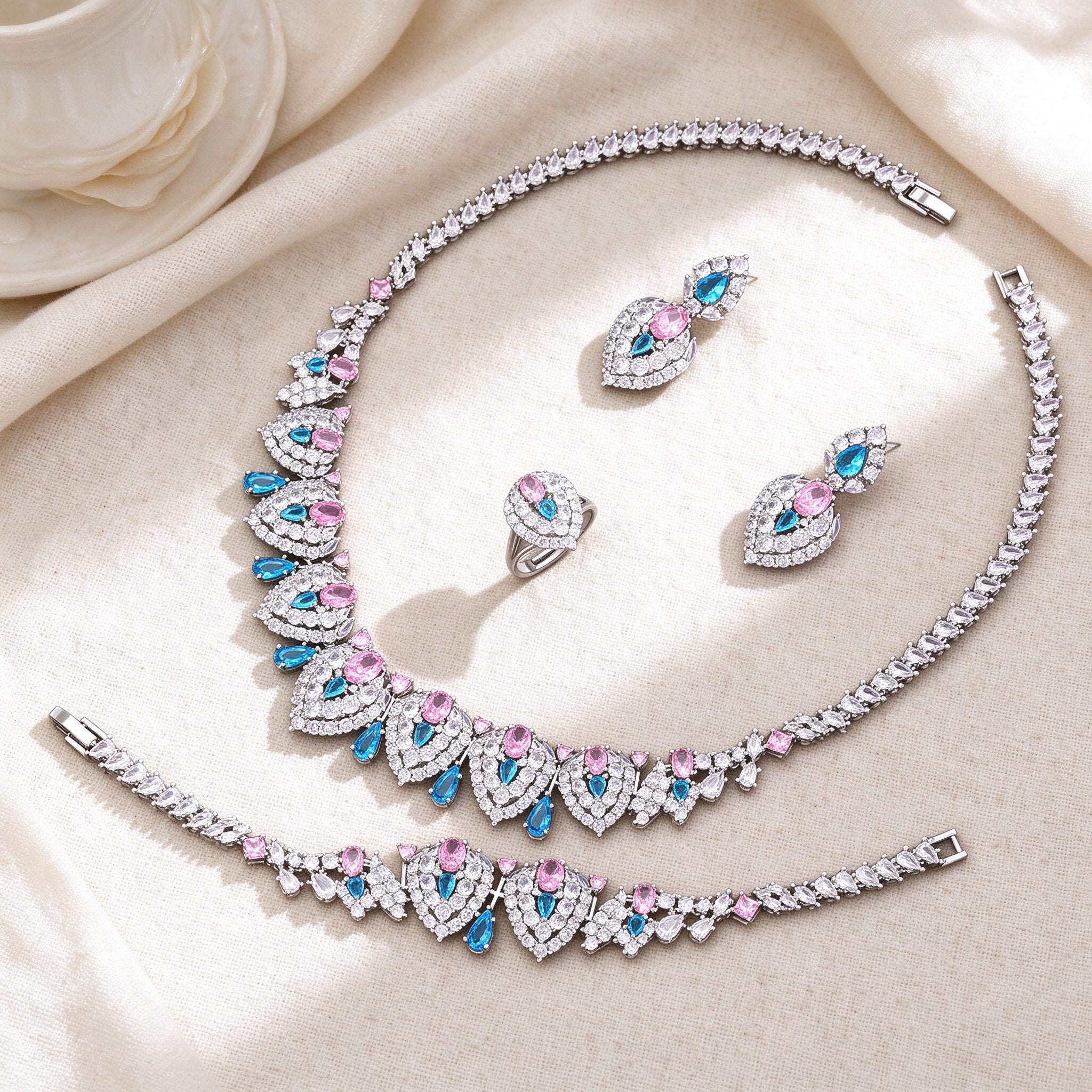 Flat lay of The Wren Wedding Suite necklace, earrings, bracelet and pink-blue cluster ring on ivory linen
