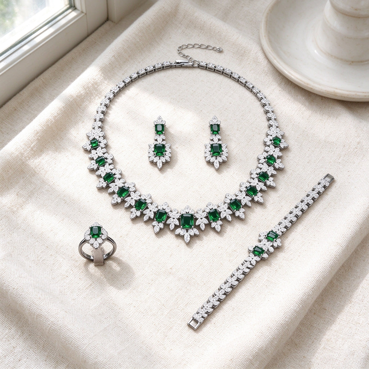 Top-down flat lay of The Twilight Bridal Parure four-piece silver emerald-green and clear CZ set on warm ivory linen