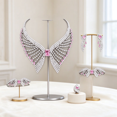 The Reagan Bridal Set — silver-tone wing-shaped bib necklace with pink emerald-cut CZ center, radiating feathered rows, earrings, bracelet and ring on an ivory background
