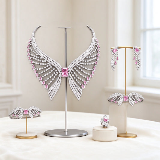 The Reagan Bridal Set — silver-tone wing-shaped bib necklace with pink emerald-cut CZ center, radiating feathered rows, earrings, bracelet and ring on an ivory background