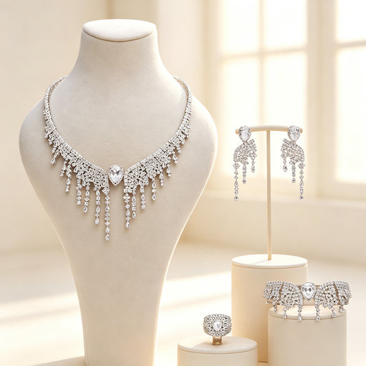 The Genevieve Bridal Set — silver-tone V-bib necklace centered on a pear CZ with cascading fringe, chandelier earrings, wide bracelet and cluster ring on ivory pedestals