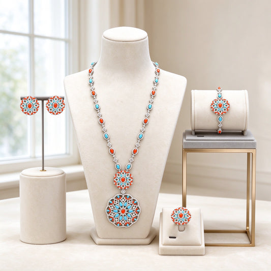 The Bud Bridal Set — silver-tone long Y-necklace alternating coral-red and turquoise-blue stones with graduated double openwork floral medallion pendant, matching stud earrings, bracelet and ring on ivory velvet