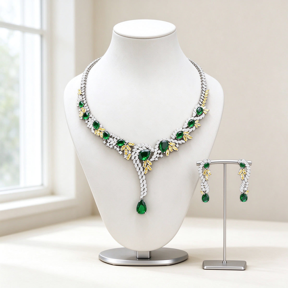 The Hadley Crystal Parure — silver-tone Y-drop lariat necklace with emerald green CZ stones, gold-tone leaf accents and matching linear earrings on an ivory background