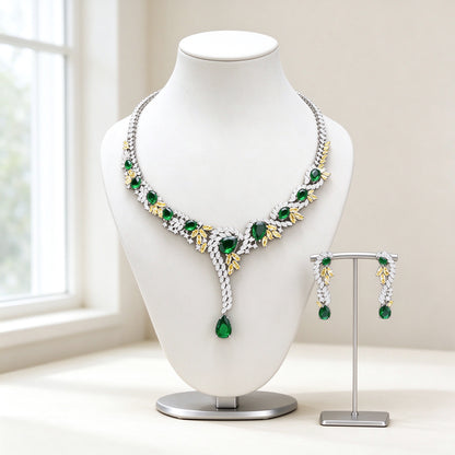 The Hadley Crystal Parure — silver-tone Y-drop lariat necklace with emerald green CZ stones, gold-tone leaf accents and matching linear earrings on an ivory background