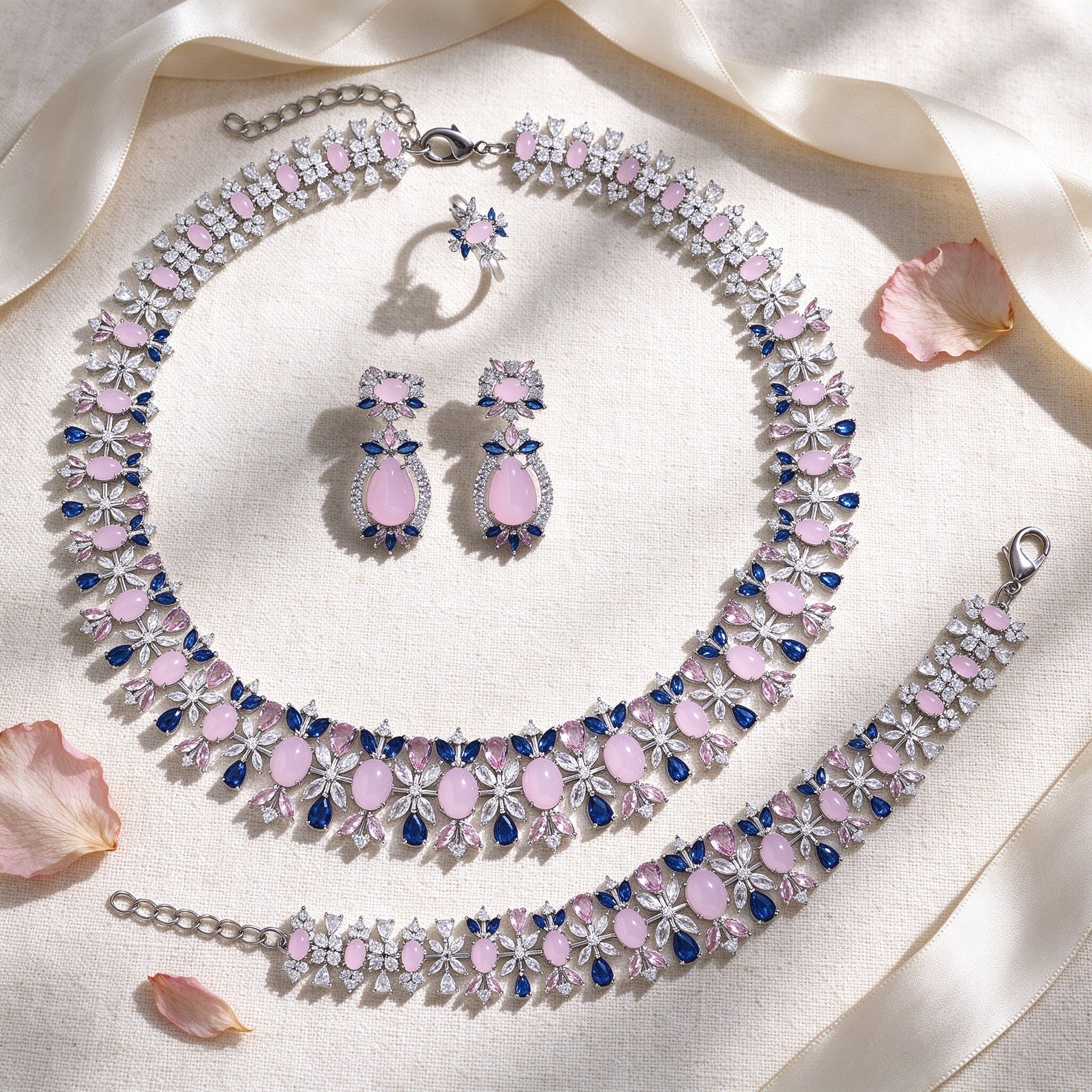 Styled flat lay of The Calliope Bridal Set necklace, earrings, bracelet and ring on cream linen with blush petals