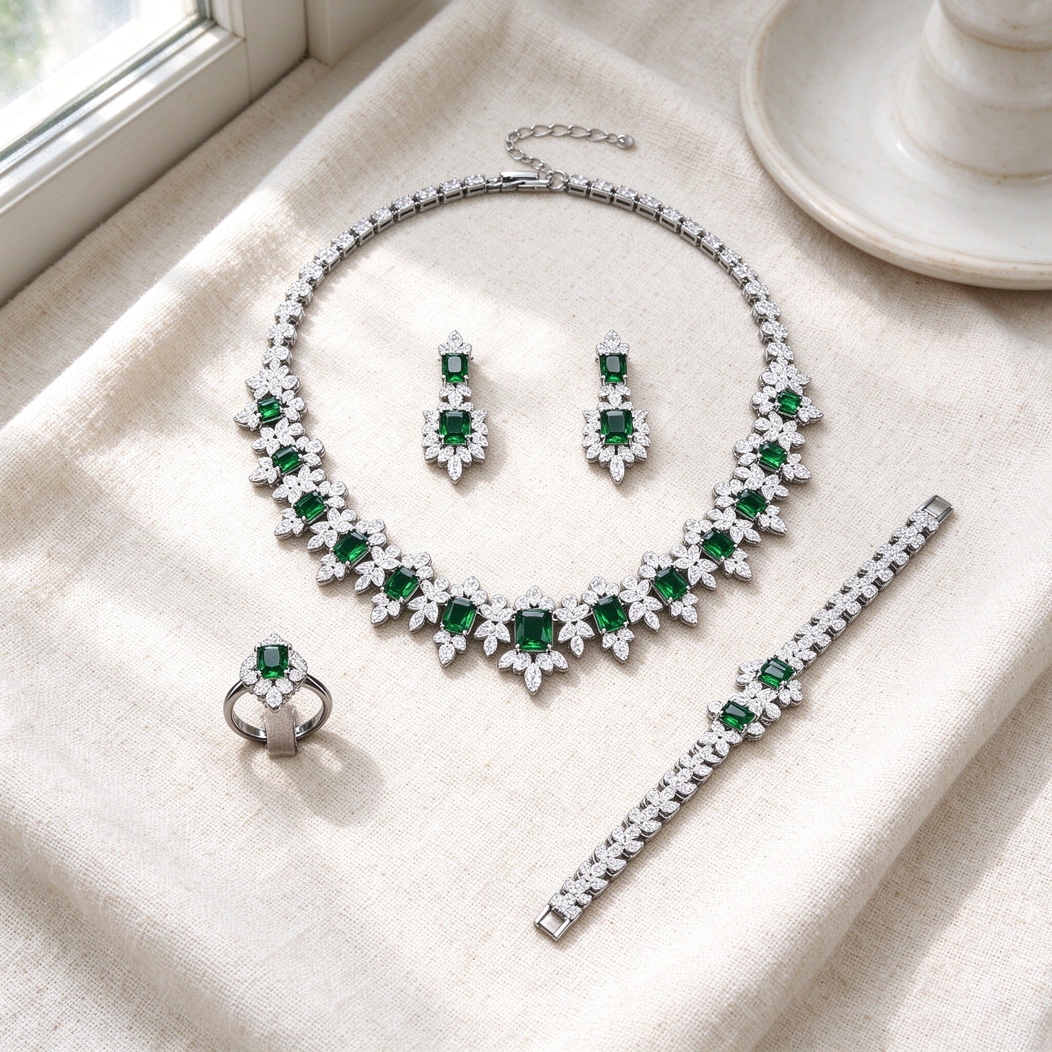 Top-down flat lay of The Twilight Bridal Parure four-piece silver emerald-green and clear CZ set on warm ivory linen